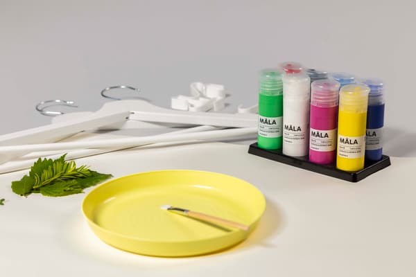 Auf einem Tisch liegen ein gelber Teller, Pinsel, Farben, Kleiderbügel und grüne Blätter.