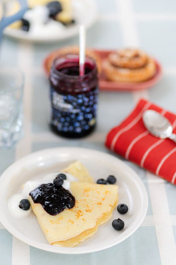 Auf einem Tisch steht ein Teller mit schwedischen Pfannkuchen, Marmelade und Preiselbeeren. Dahinter ist ein Glas Marmelade zu erkennen.