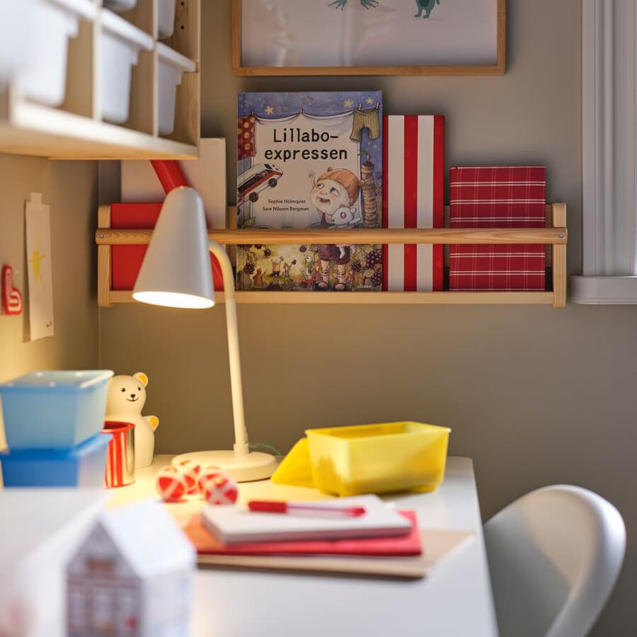 Auf einem Tisch steht eine Lampe die auf Zettel leuchtet. Daneben hängen Regale mit Boxen und bunten Büchern. Vor dem Tisch steht ein weißer Stuhl.