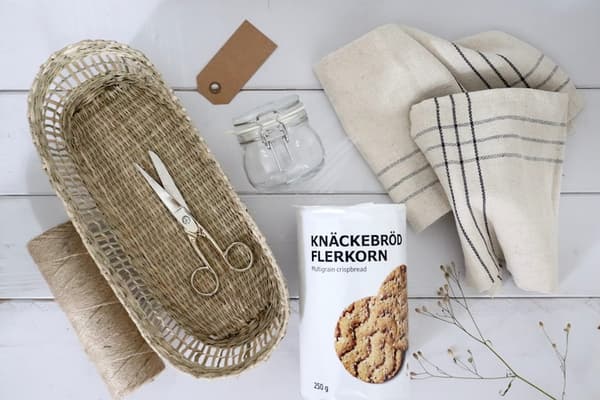 Auf einem weißen Holztisch liegen ein kleines, längliches PENNFISK Brotkörbchen aus Naturfasern, darin eine Schere. Daneben befinden sich Naturfaden, eine Packung Knäckebrot, ein Einmachglas, VÅRARV Servietten und ein Geschenkanhänger.