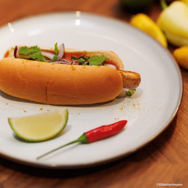 Auf einem weißen Teller liegt ein Hotdog. Daneben liegt eine Limete und eine kleine Chili.