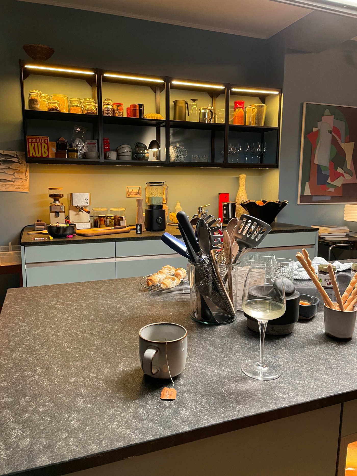 Auf einer Kücheninsel stehen mehrere Utensilien in einem Utensilienständer, eine Teetasse mit Teebeutel darin, in Weinglas und verschiedene Snacks.