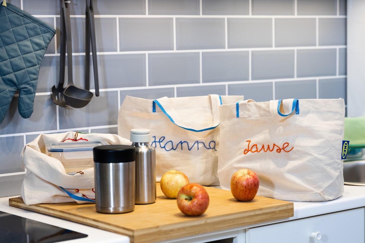 Auf einer Küchenplatte stehen 2 weiße Taschen und davor liegen 3 rote Äpfel.