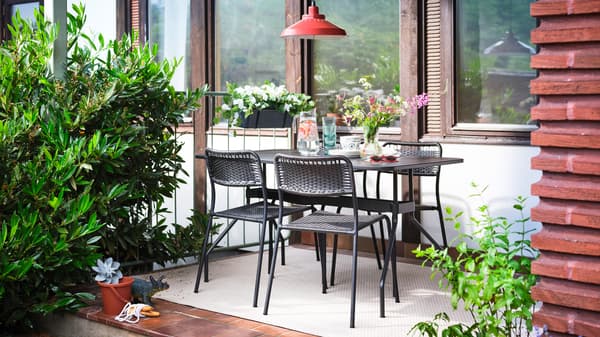 Auf einer Terrasse stehen ein VIHOLMEN Tisch und vier Stühle auf einem cremefarbenen Teppich, u. a. mit einer Vase Blumen, einer Kanne Wasser und Gläsern.