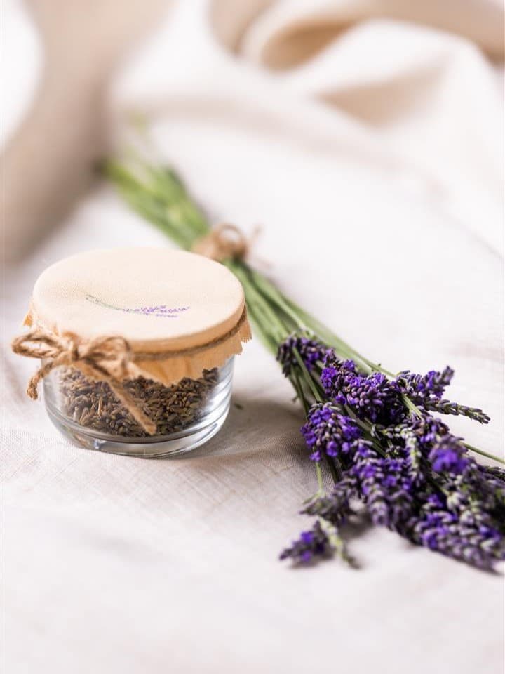 Auf einer weißen Unterlage liegt ein Glas mit Stoffdeckel und ein Ast von Lavendel.