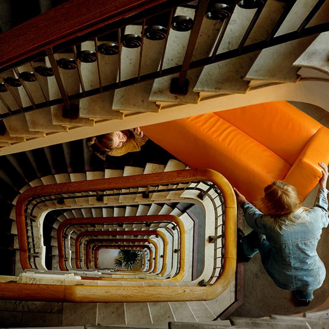 Aus der Vogelperspektive sieht man einen Stiegenaufgang hinanb und zwei Personen tragen ein gelbes Sofa auf den Stiegen