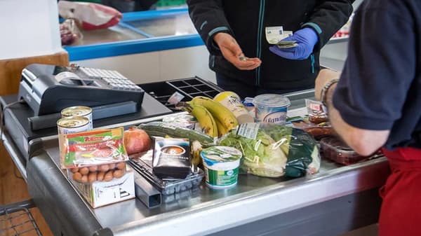 Ausgabe von Lebensmitteln an einem Verkaufstresen eines Tafelladens: Auf dem Tresen liegen Obst, Gemüse, Konserven, Bananen, Eier und verpackte Waren. Zwei Personen stehen sich gegenüber, eine Person zählt Bargeld, die andere reicht Geld entgegen. Im Hintergrund ist eine Kasse zu sehen.