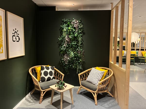 Avskild sittgrupp i IKEA Malmös personalmatsal med rottingfåtöljer, mörkgrön vägg, växtinstallation och trädetaljer som skapar en lugn och ombonad pausmiljö.