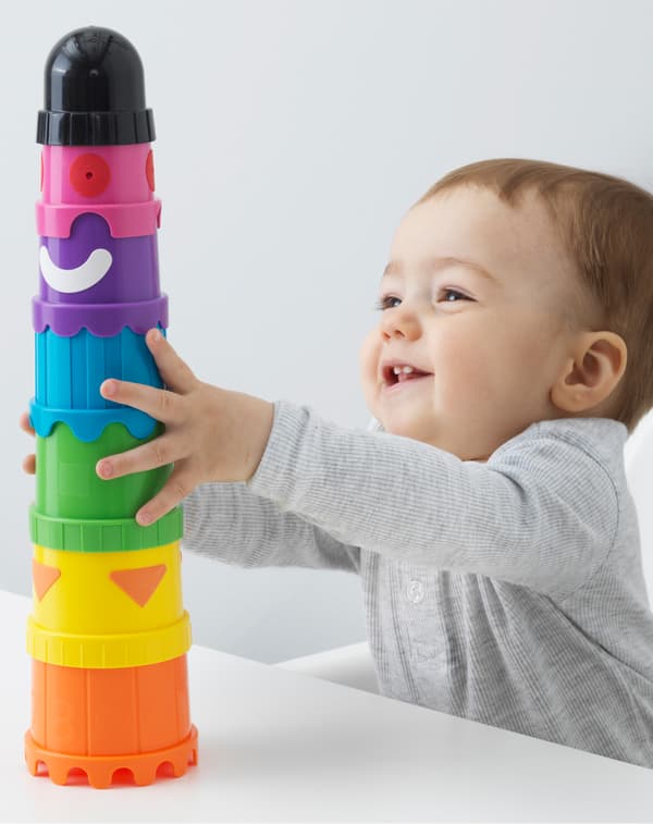 Baby holding onto a tower of MULA bright coloured cups that stack together at a table while sitting in a highchair.