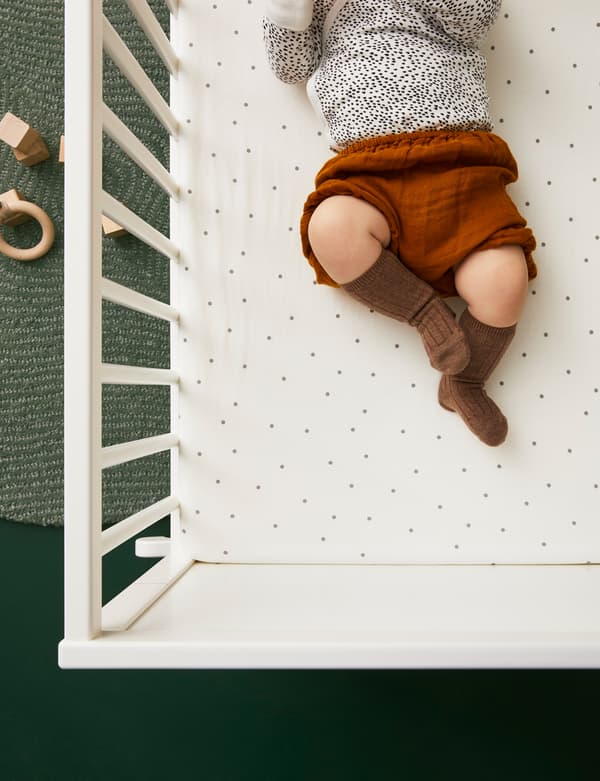 Baby lying in a cot with LENAST fitted sheet dressed in cute clothes with wooden toys on a rug on the floor nearby.
