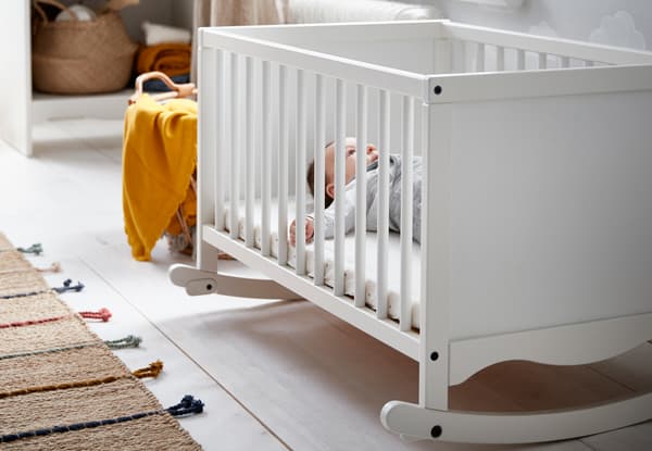 Baby lying in an IKEA SOLGUL cot in a room with white wooden floorboards and natural textile rug and basket.