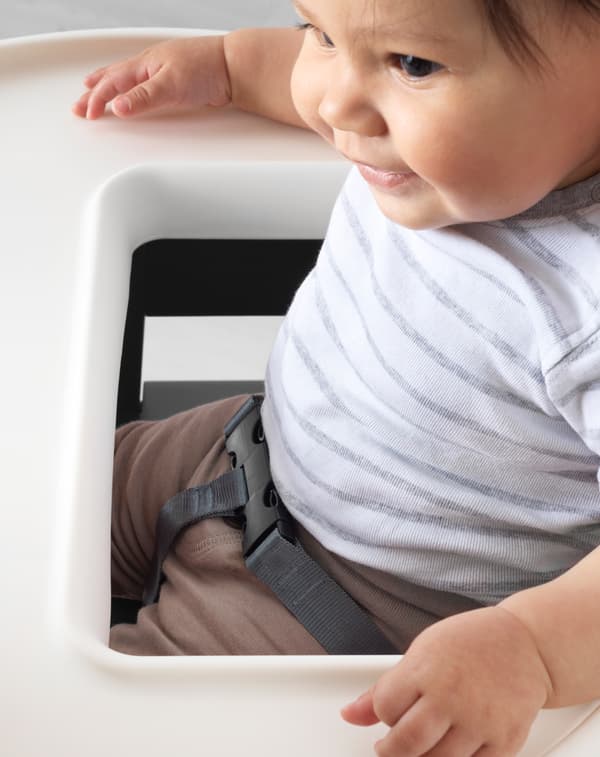 Baby sitting in IKEA ANTILOP highchair with safety belt on and arms on the tray table smiling.