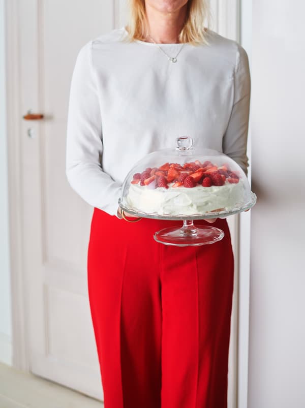Badan dan kaki seorang wanita yang berpakaian untuk ke parti sedang memegang bekas hidangan berpenutup kaca ARV BRÖLLOP yang mengandungi kek strawberi.