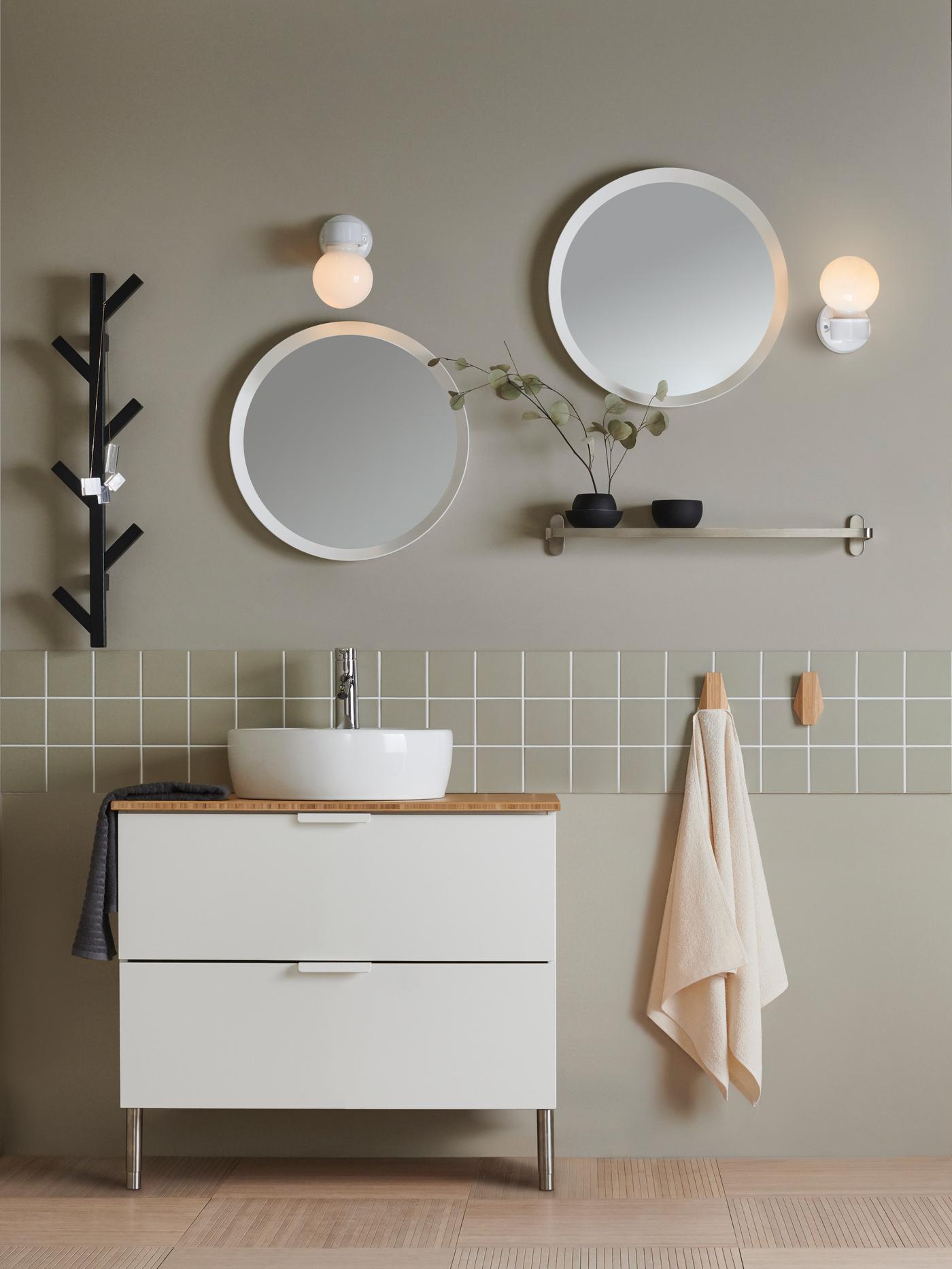 Badrumsinteriör med två runda speglar, vägglampor, vit kommod, växter och handdukar på handdukskrokar.
