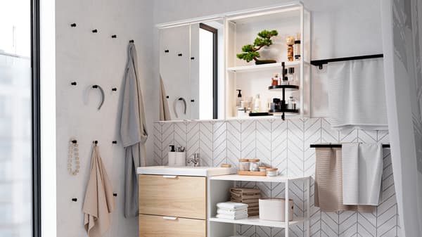 Bagno con mobili bianchi, ganci neri sulla parete, scaffale con asciugamani, profumi, un bonsai e un accappatoio.