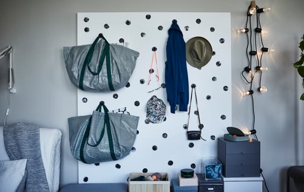 Bags and hats hanging on a white wall storage with black dots, made of a painted plywood sheet and kitchen knobs.