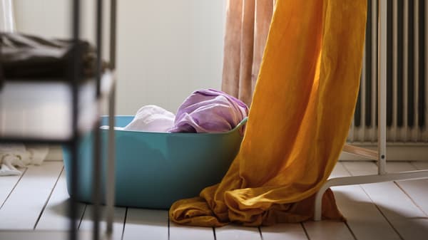 Bakul kain cucian TORKIS berwarna biru dengan tekstil panjang yang berwarna-warni di dalamnya dan juga tergantung pada rak pakaian MULIG di sebelahnya.