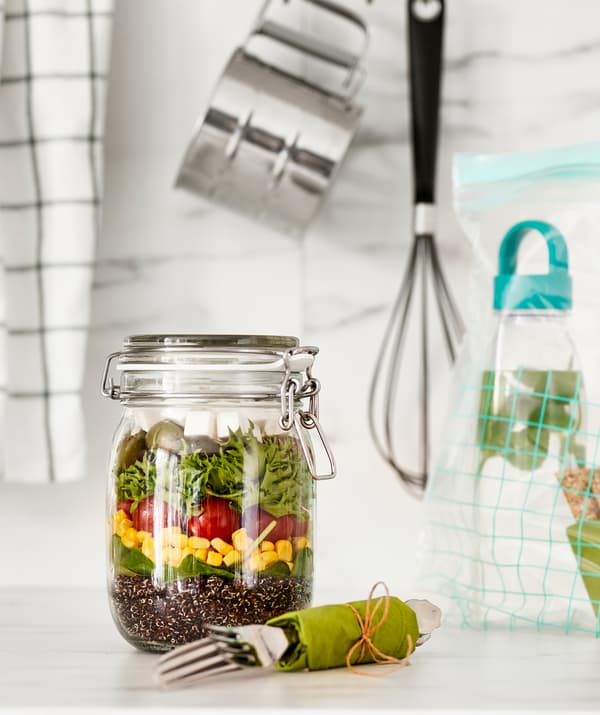 Balang kaca yang tinggi dengan penutup terpasang, mengandungi salad gaya Greek yang berlapis; kit perkakas makan dan botol air di tepinya.
