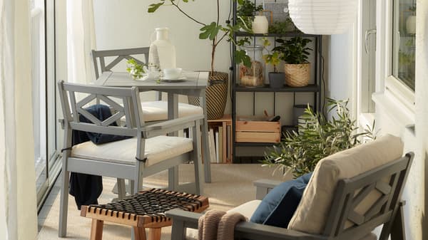 Balcon clos avec meuble BONDHOLMEN gris, suspensions rondes, tapis beige avec rideaux blancs et plusieurs plantes.