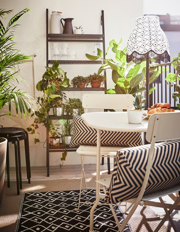 Balcon décoré avec une petite table, un lampadaire, deux tapis, une étagère, des plantes et des tabourets supplémentaires.