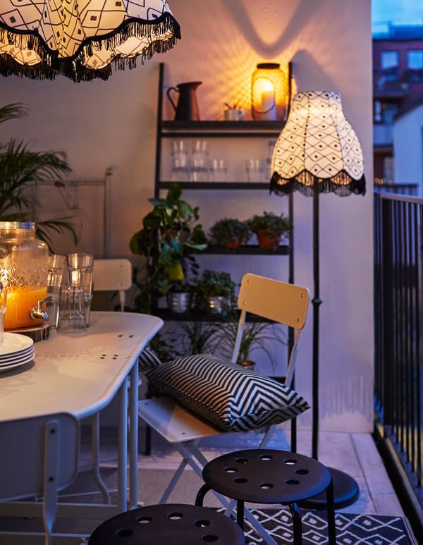 Balcon en soirée, avec table déployée et dressée pour le repas, lampadaire, textiles, étagère, plantes vertes et tabourets supplémentaires.