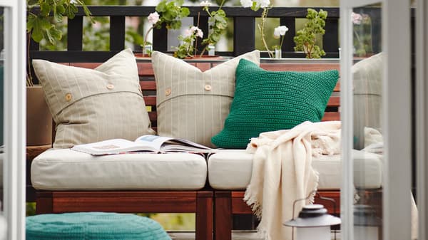 Balcone con divano da esterno in legno con cuscini bianchi e verdi, un plaid e un libro aperto, e piante sullo sfondo.