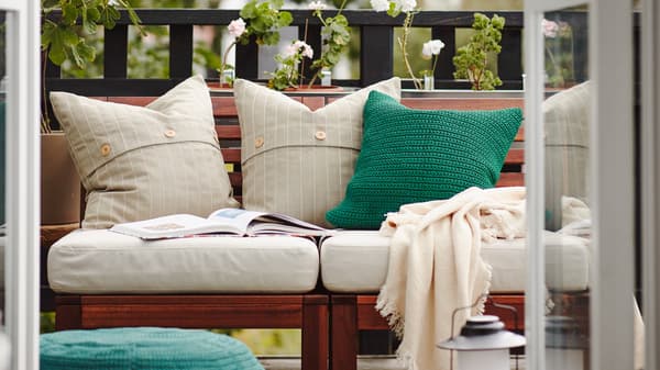Balcone con divano da esterno in legno con cuscini bianchi e verdi, un plaid e un libro aperto, e piante sullo sfondo.