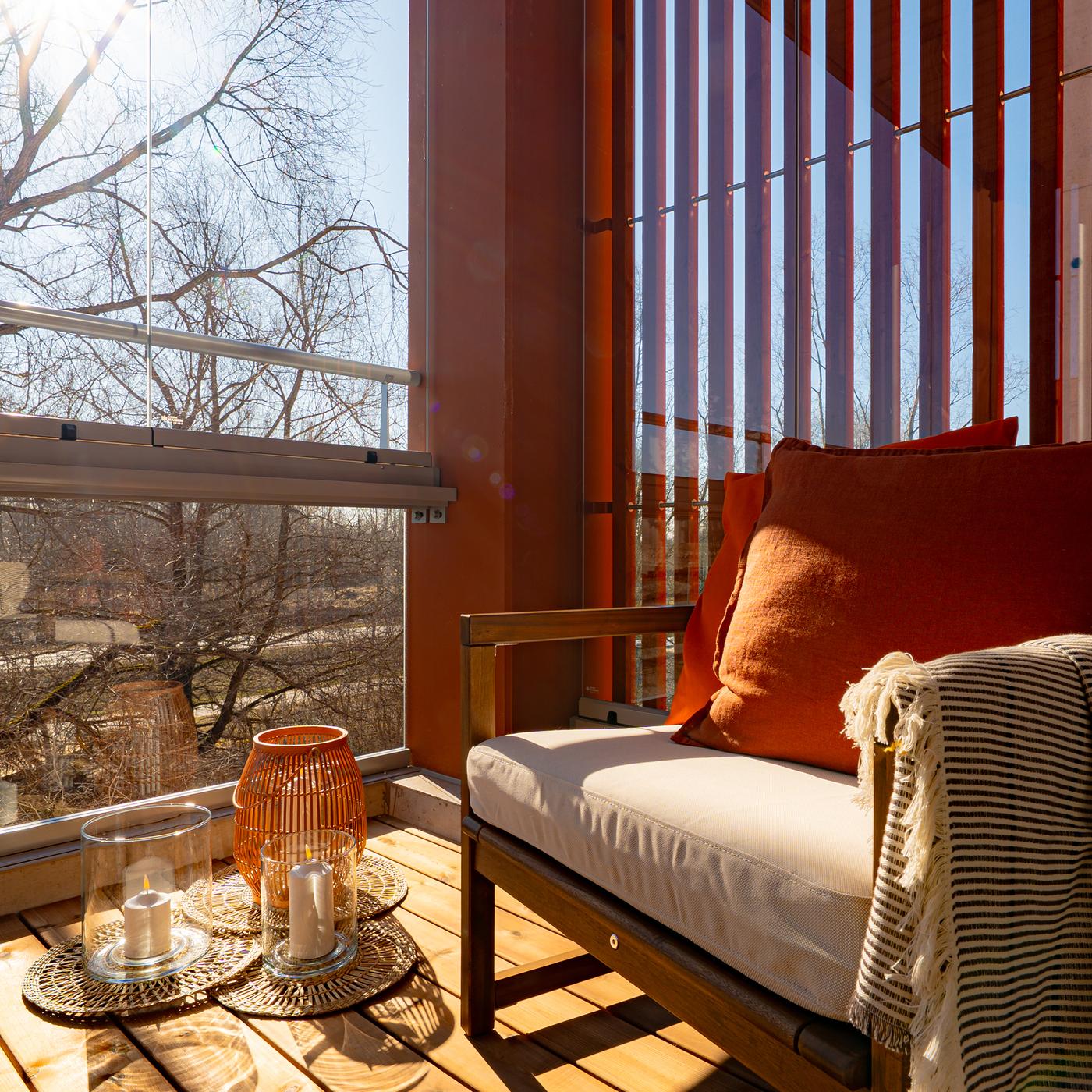 Balcony seating area with a wooden armchair, white and terracotta cushions, glass candle holders, and wooden decking overlooking trees.