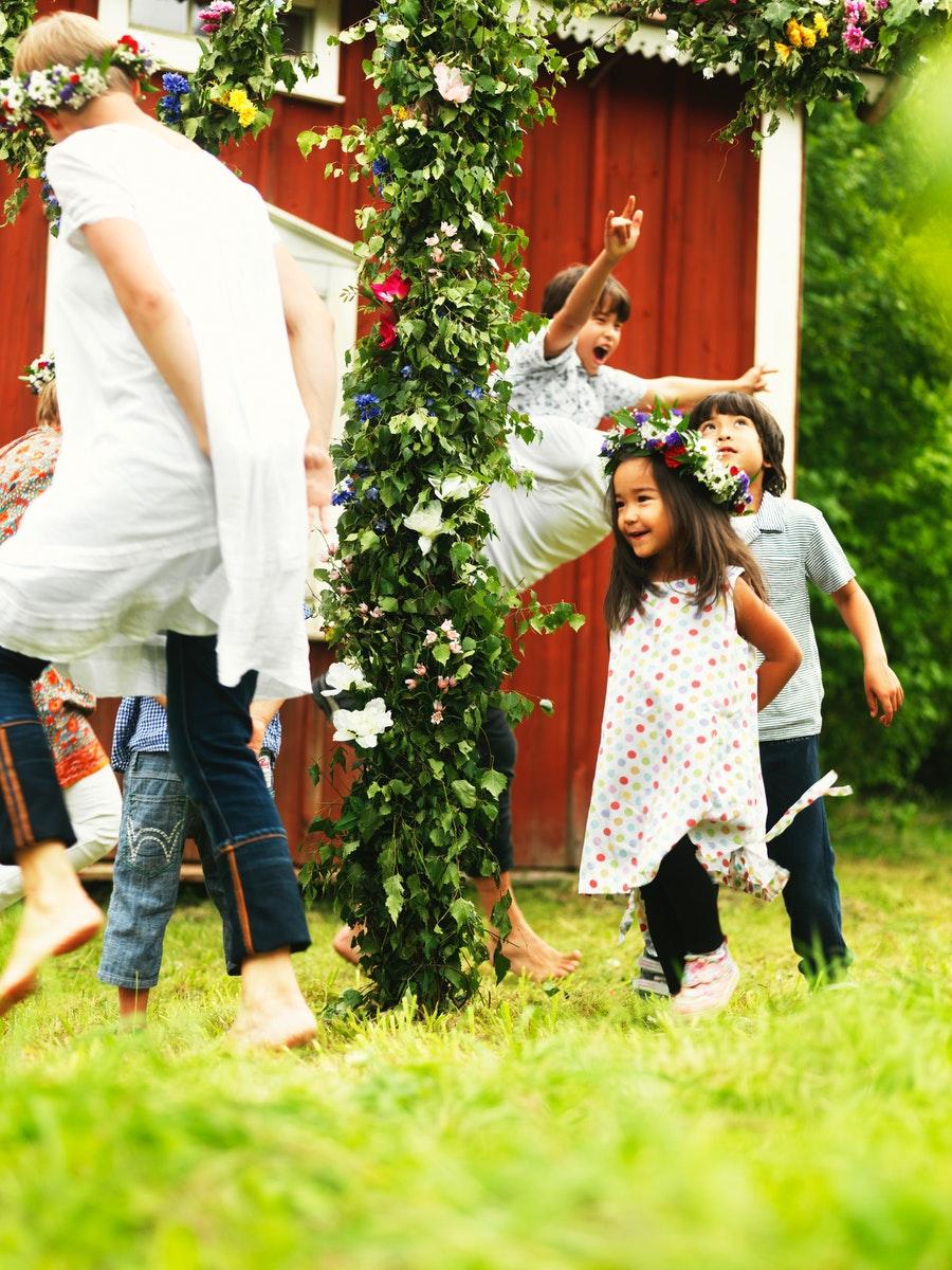 Bambini e giovani in giardino stanno giocando a rincorrersi mentre indossano delle coroncine di fiori colorati in occasione della festa del Midsommar.