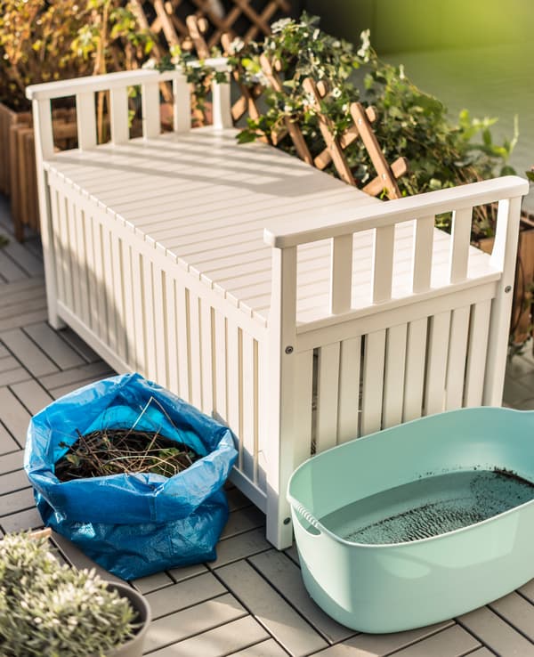 Banc de rangement blanc, bassine en plastique remplie d’eau de pluie et sac IKEA bleu rempli de compost, au jardin.