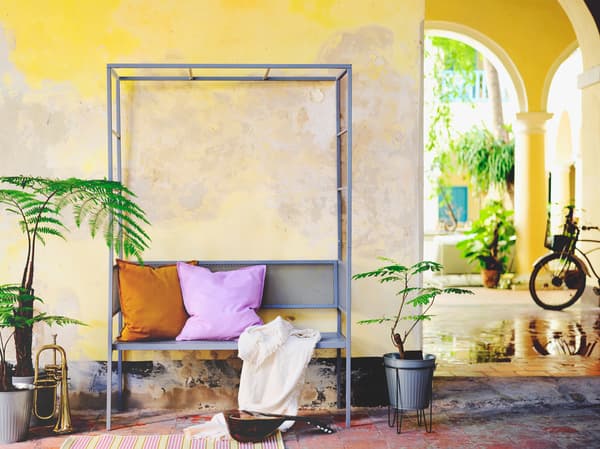 Banco con pérgola colocado fronte a unha parede amarela complementado con coxíns en cor púrpura e laranxa.