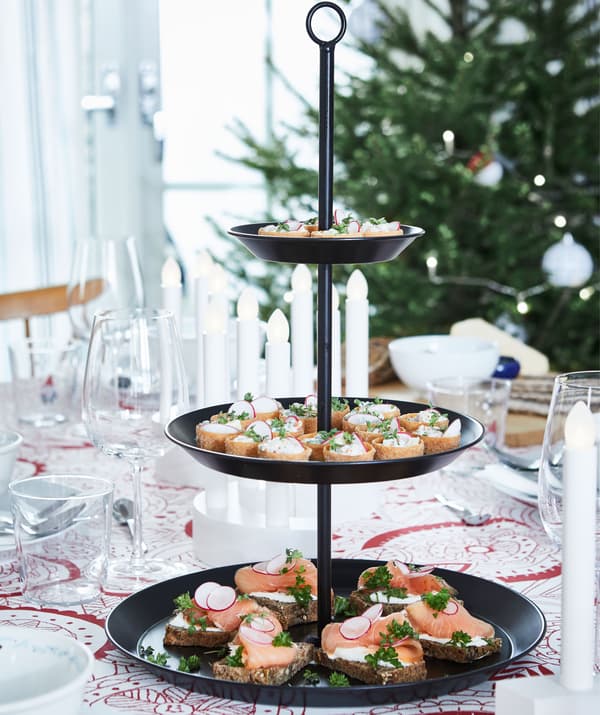 Bandexa de tres pisos con elaborados petiscos encol dunha mesa preparada para unha celebración. Árbore de Nadal no fondo.