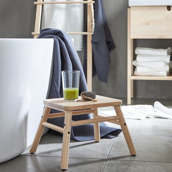 Bangku tangga VILTO yang dibuat daripada kayu birch terletak di sebelah tab mandi dengan segelas smoothie hijau di atasnya di dalam bilik mandi minimalistik.