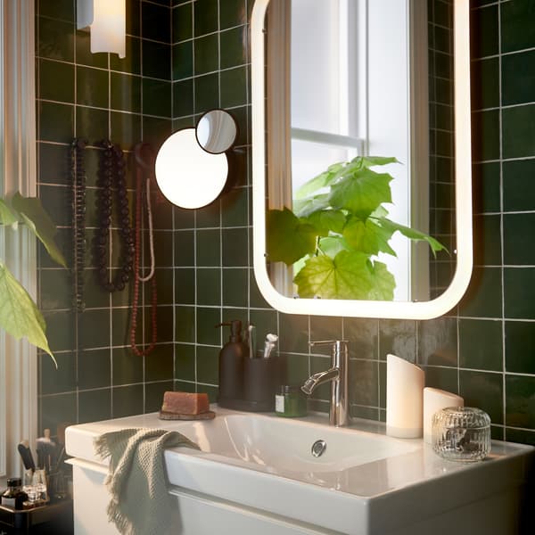 Baño con un espejo blanco con iluminación integrada sobre un mueble de lavabo, además de dos velas gruesas LED y una planta.