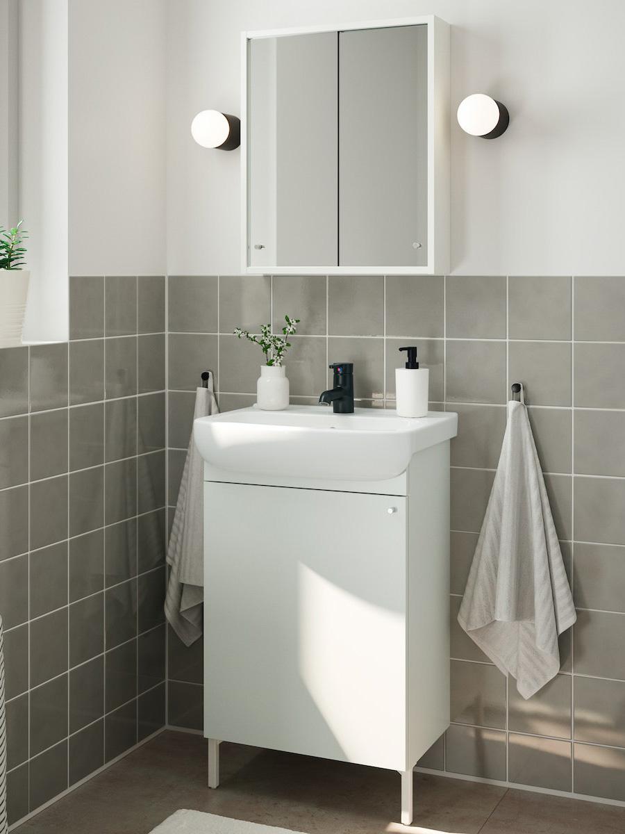 Bany petit amb moble blanc de lavabo, mirall amb armariet, dues tovalloles penjades i parets enrajolades en to gris clar.