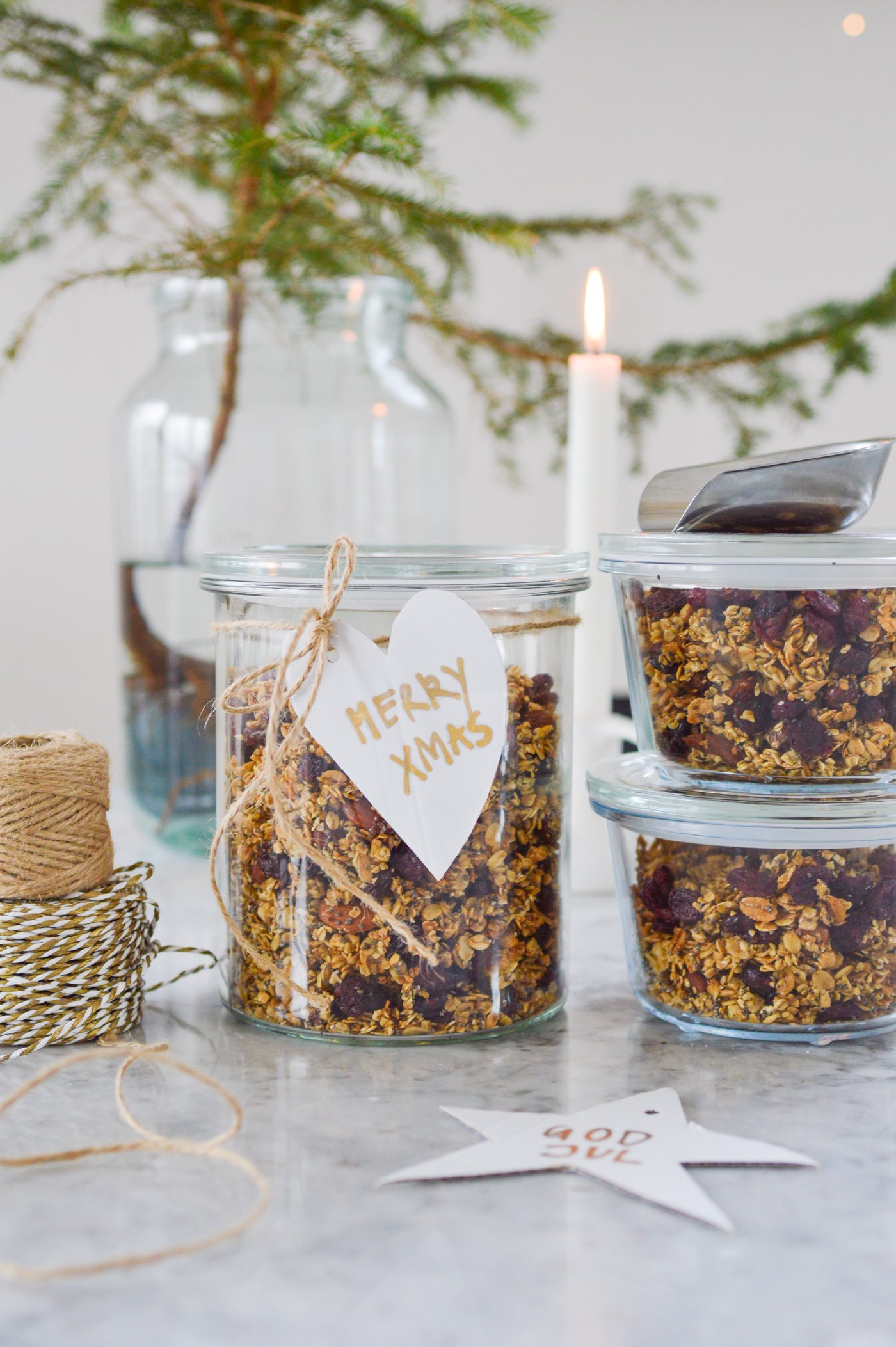 Barattoli di cereali con un'etichetta "Merry Xmas" sono disposti su un vassoio di marmo, circondati da cordoncini, una candela bianca e un piccolo albero, creando un'atmosfera festosa e casalinga.