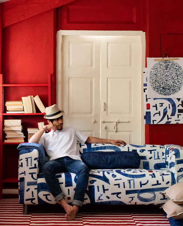 Barefoot man in Panama hat sitting on blue and white sofa in a room with red walls, bookshelf, white door and two-tone rug.