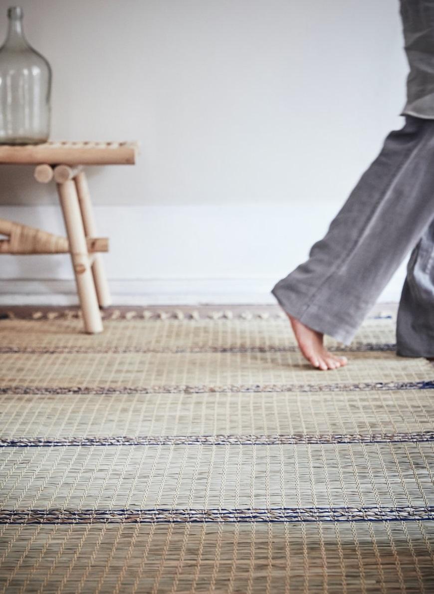 Barefoot person stepping onto a textured woven rug in a minimal, neutral-toned room with wooden furniture and soft lighting.