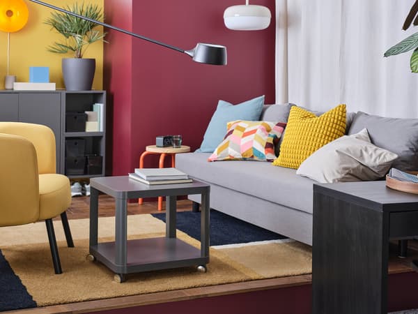 Barevný obývací pokoj zařízený v retro stylu, se žlutým křeslem, šedou pohovkou, různobarevnými polštáři a šedými skříňkami EKET.