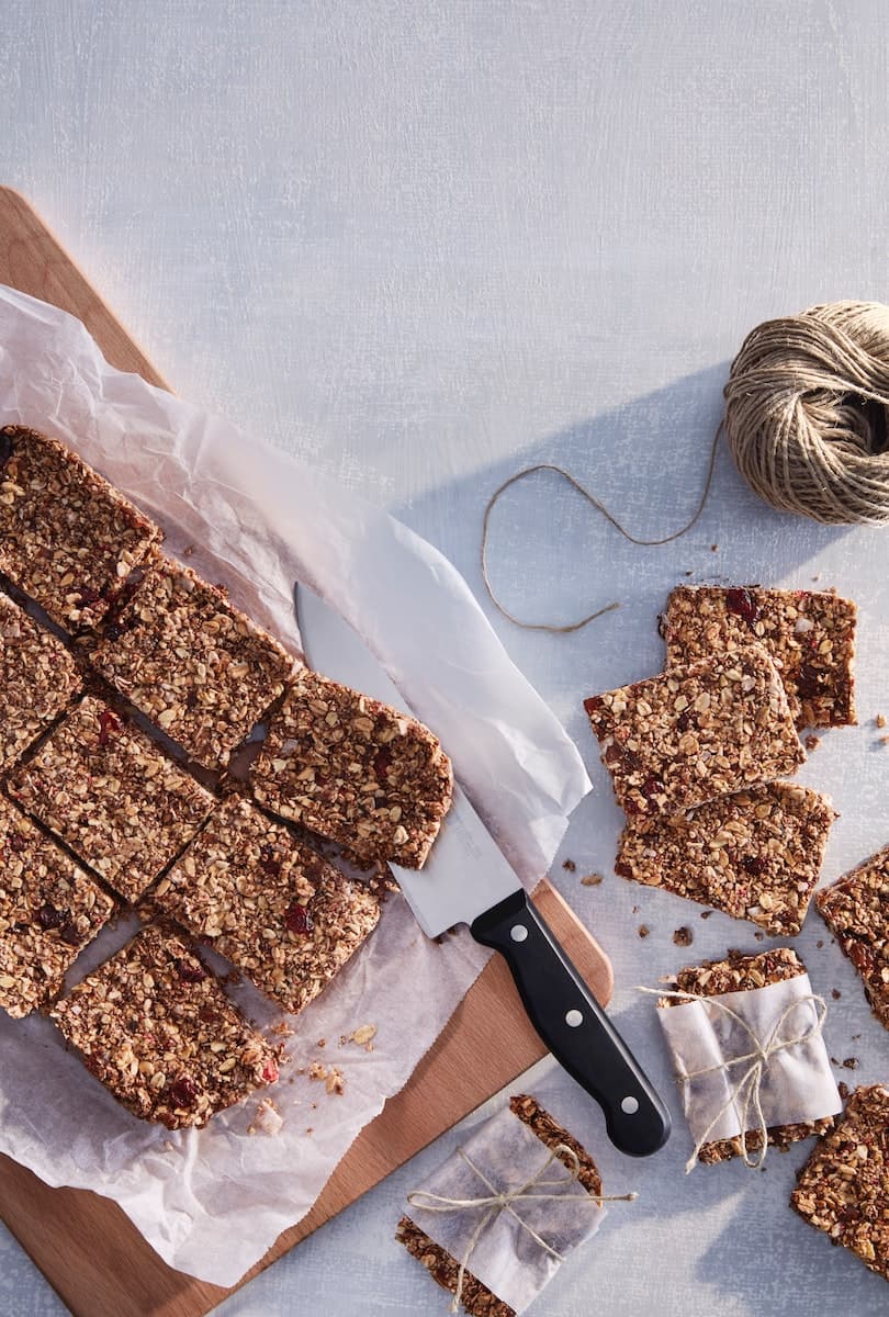Barras de granola cortadas en forma rectangular sobre un papel de hornear blanco y una tabla de cortar de madera, junto a un cuchillo de cocina y dos trozos envueltos.