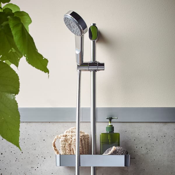 Barre de douche chromée BROGRUND avec douchette, fixée au mur. Elle est dotée d’un plateau pouvant accueillir une éponge et un distributeur de savon.