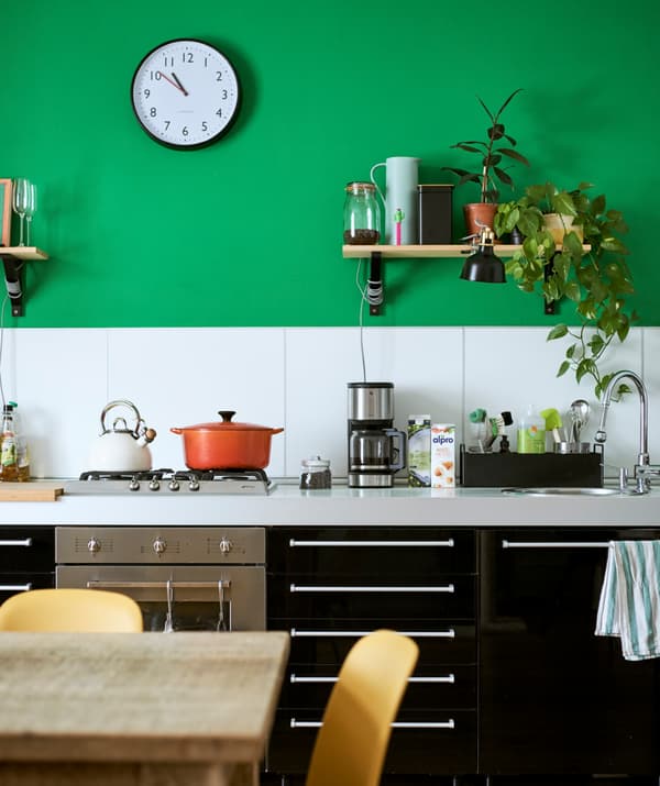 Basi da cucina con ante e cassetti in nero lucido sullo sfondo di una parete verde con piastrelle bianche. Uno scaffale con piante, contenitori e una lampada nera con morsetto.