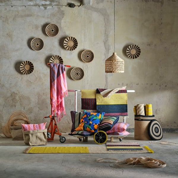 Baskets made of natural materials, patterned cushion covers, handwoven wall decorations, a lampshade and some rugs from the MÄVINN collection have been arranged in a room with concrete walls and floors.