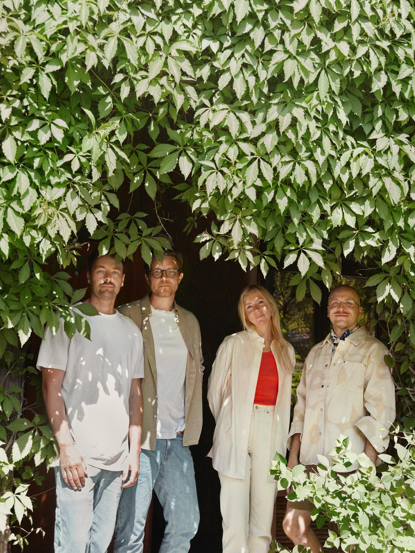 BASTUA 바스투아 컬렉션의 디자이너 네 명이 밝은 색감의 캐주얼한 옷차림을 하고 잎이 무성한 잎사귀 아래 서 있는 모습.