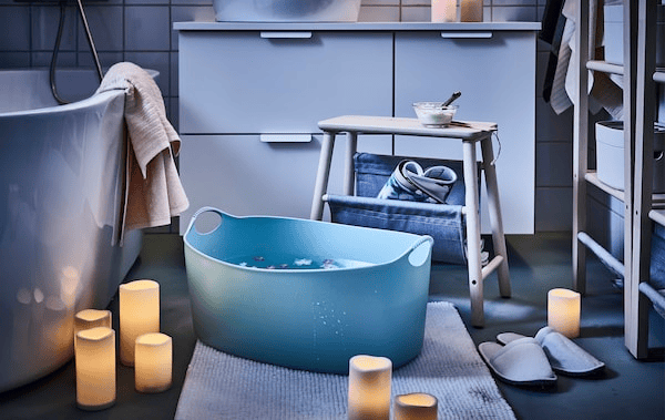 Bathroom mood-lit by scattered LED block candles; a stool placed next to a big footbath, flowers floating in it.