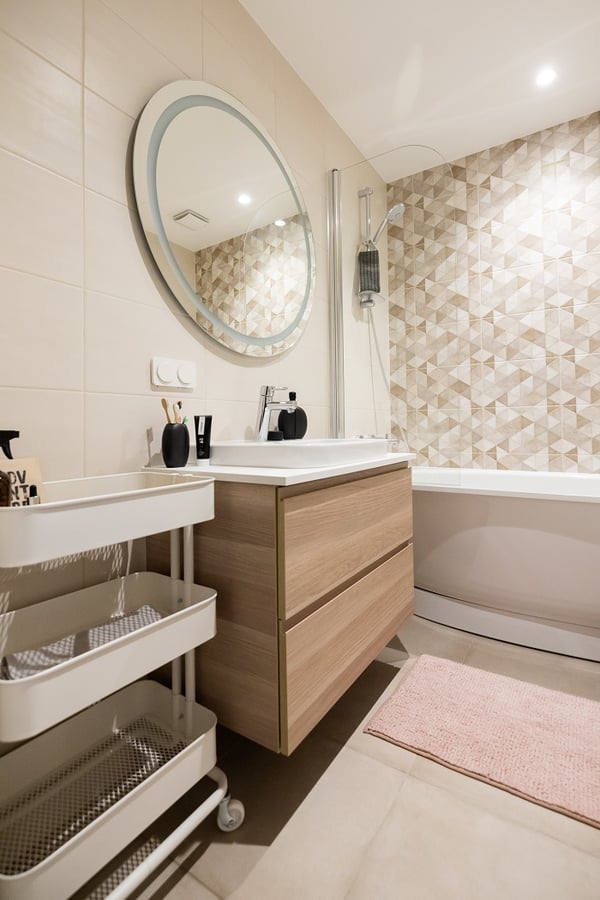 Bathroom with round mirror, wooden vanity, geometric tile wall, and pink bath mat.