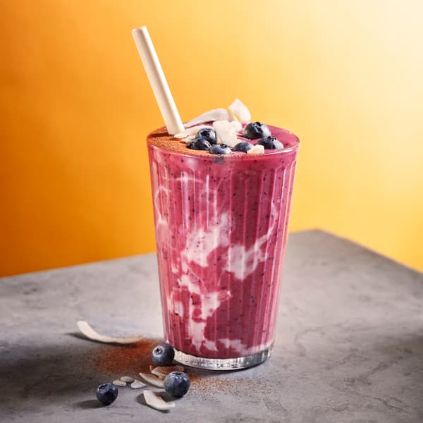 Batido de mirtilo num copo alto colocado numa mesa cinzenta em frente a uma parede laranja.