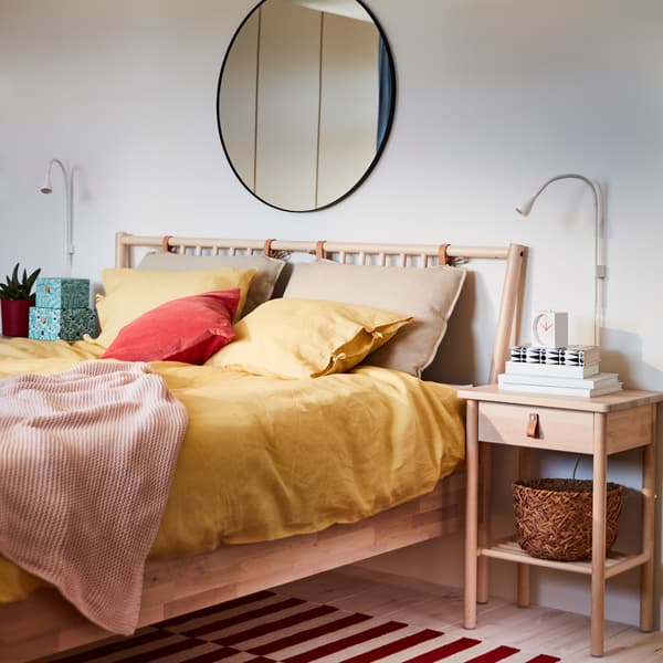Bed frame and bedside table in birch with tanned leather details, a round mirror, a pale pink throw and yellow bed textiles.