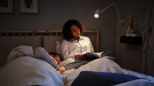 ベッドに座り読書ランプの下で本を読む女性と、隣で寝ている男性。