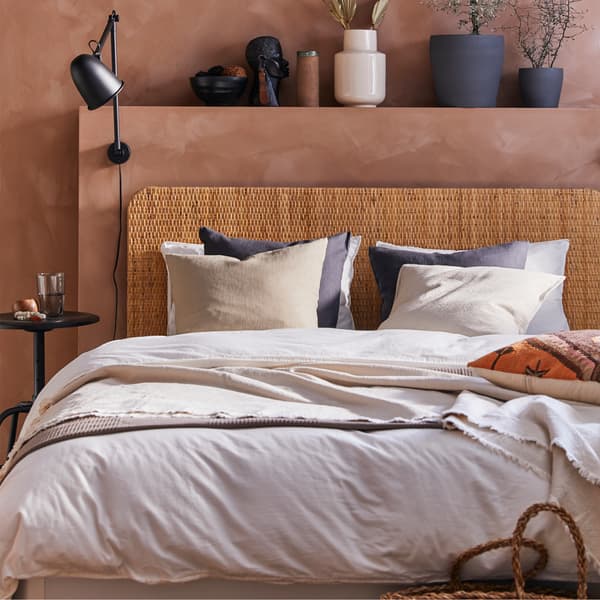 Bedroom with a bed with rattan headboard, dark gray plant pots, black wall lamp, white bed textiles and beige cushion covers.